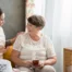 Smiling caregiver with her hand on an older woman’s shoulder as they share tea on the couch, showing home care vs assisted living