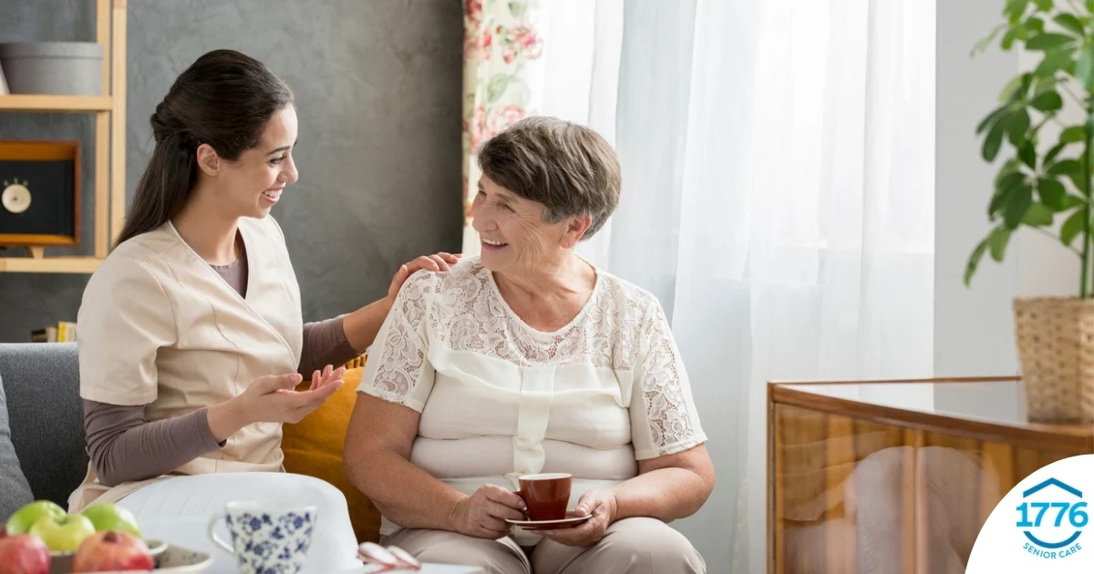 Smiling caregiver with her hand on an older woman’s shoulder as they share tea on the couch, showing home care vs assisted living